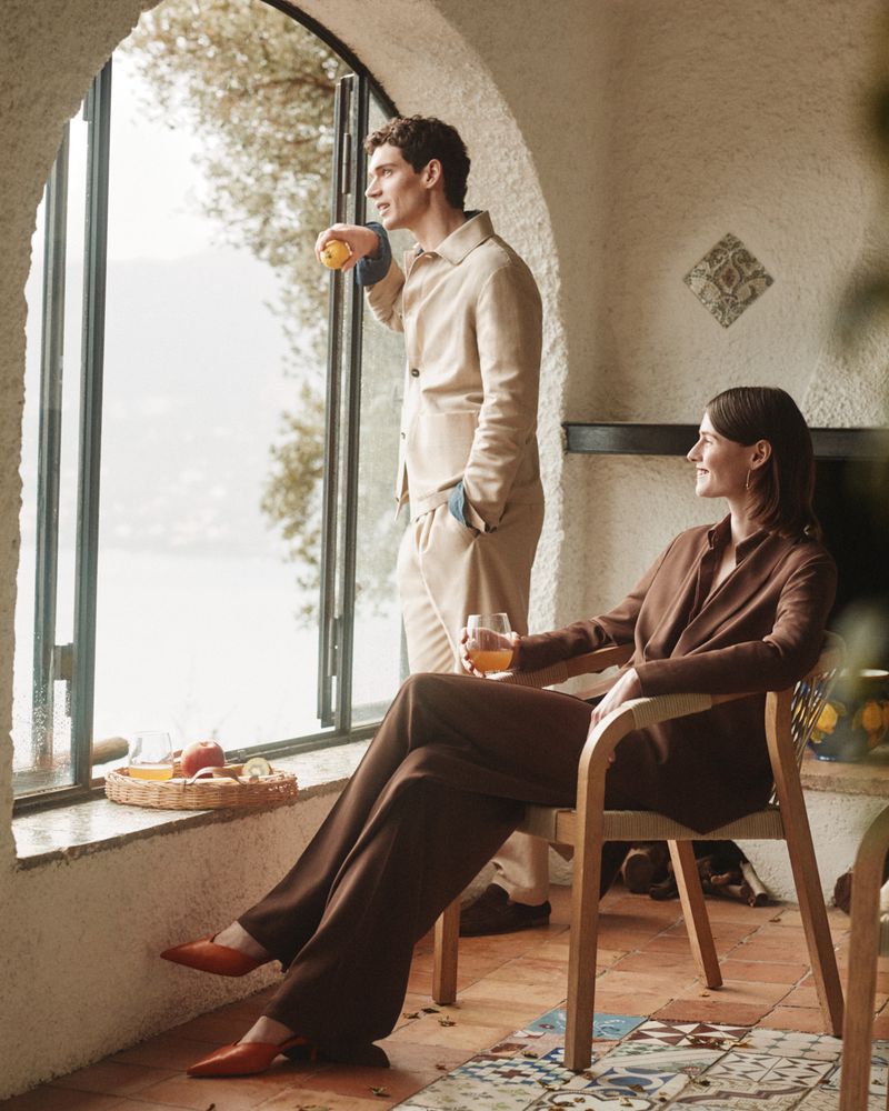 Man standing by a window holding fruit and a woman seated in a chair holding a glass of juice inside a rustic interior for Slowear SS26
