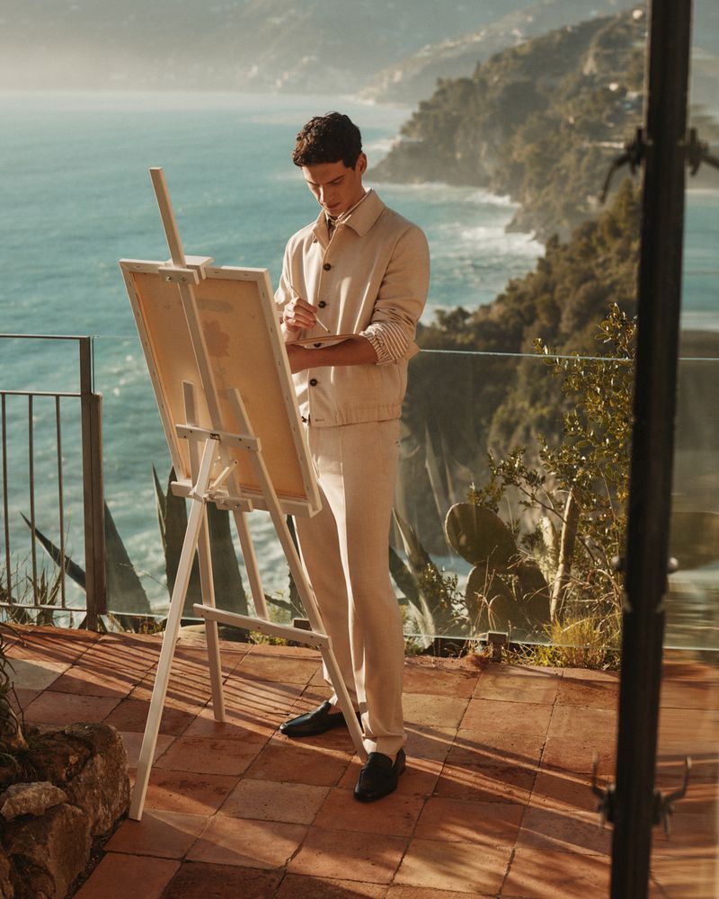 A man in casual attire paints on a canvas with ocean and coastal mountains in the background on a sunny day outdoors.