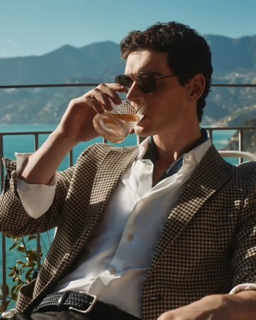 A stylish man in sunglasses and a blazer sips a drink on a balcony overlooking a scenic mountain and ocean view on a sunny day.