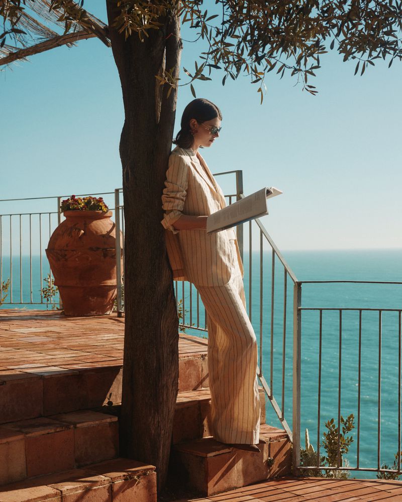 A young woman in a tan striped suit stands on a red-brick patio overlooking the ocean, reading a newspaper on a sunny day.