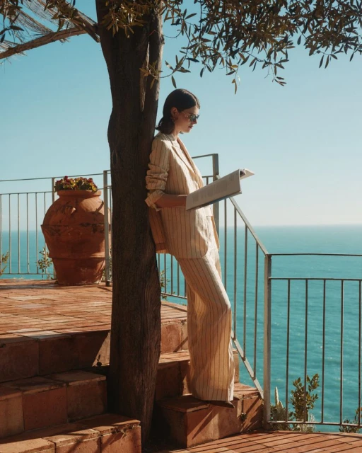 A young woman in a tan striped suit stands on a red-brick patio overlooking the ocean, reading a newspaper on a sunny day.