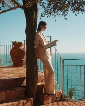 A young woman in a tan striped suit stands on a red-brick patio overlooking the ocean, reading a newspaper on a sunny day.