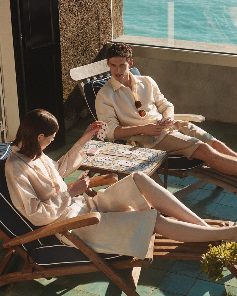 A casually dressed couple plays cards on a sunlit balcony overlooking the ocean, evoking a relaxed and leisurely summertime mood.