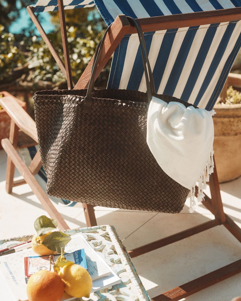Woven leather tote bag hanging on a striped deck chair, lemons on a table, outdoor setting with natural light. Fashion and travel.