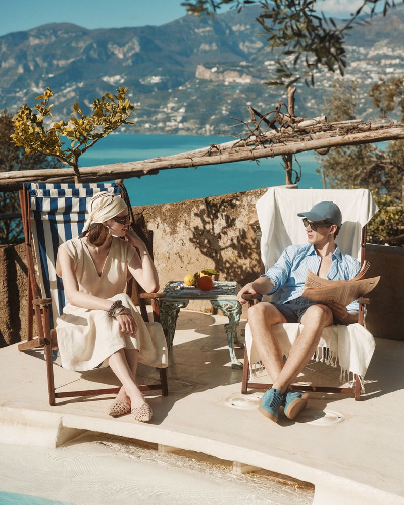 A couple lounges outdoors near the water; a man reads a newspaper while a woman sits relaxed in a beach chair, with blue water backdrop.