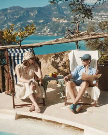 A couple lounges outdoors near the water; a man reads a newspaper while a woman sits relaxed in a beach chair, with blue water backdrop.