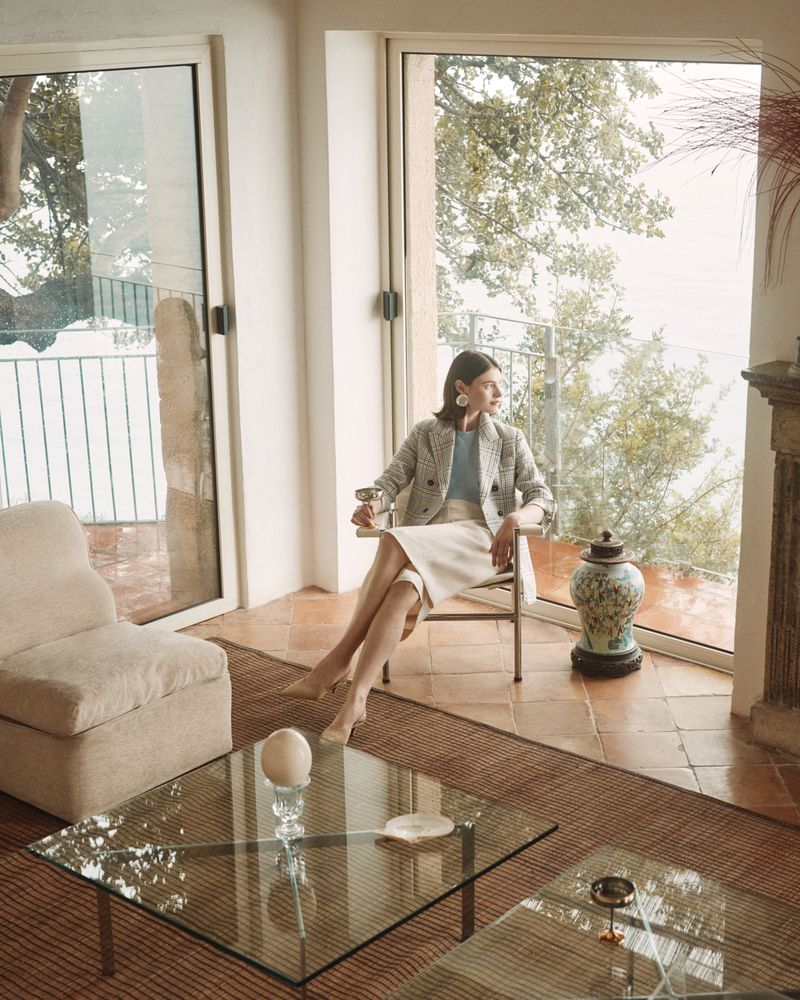 Adult woman sits in a chair by a window in an indoor living space with glass coffee tables and a view of trees and greenery outdoors.