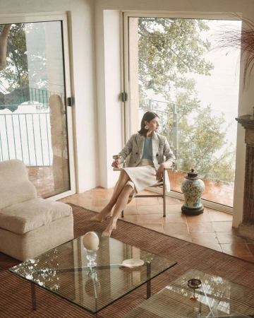 Adult woman sits in a chair by a window in an indoor living space with glass coffee tables and a view of trees and greenery outdoors.