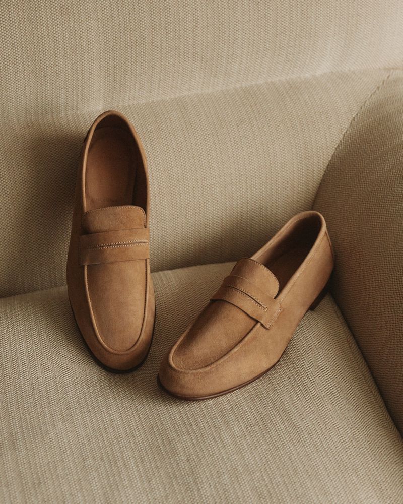 Two tan suede penny loafers sit on a light beige textured sofa cushion in a softly lit, close-up product shot.