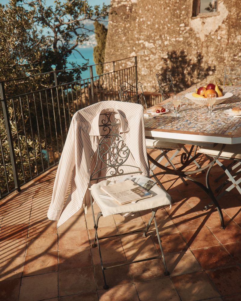 Outdoor still life with a shirt draped over a wrought iron chair, a table set with fruit, glassware, and a newspaper on the chair seat.