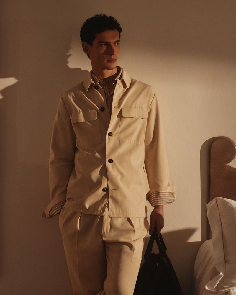 A young adult man in a tan button-down shirt and pants leans against a beige wall indoors, holding a black tote bag.