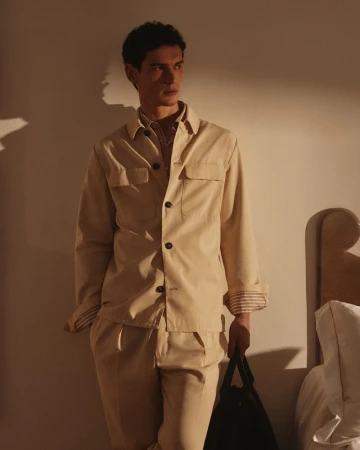 A young adult man in a tan button-down shirt and pants leans against a beige wall indoors, holding a black tote bag.