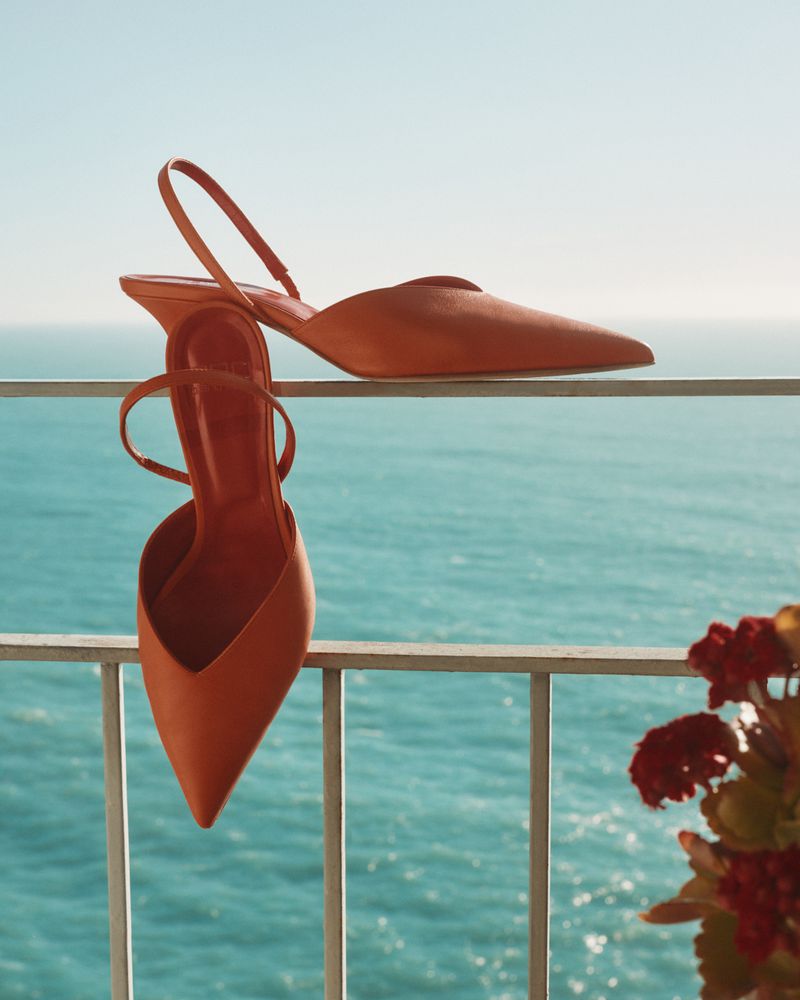 Elegant slingback heeled shoes, in warm orange hue, are displayed against a backdrop of serene blue sea and sky, promoting fashion and style.