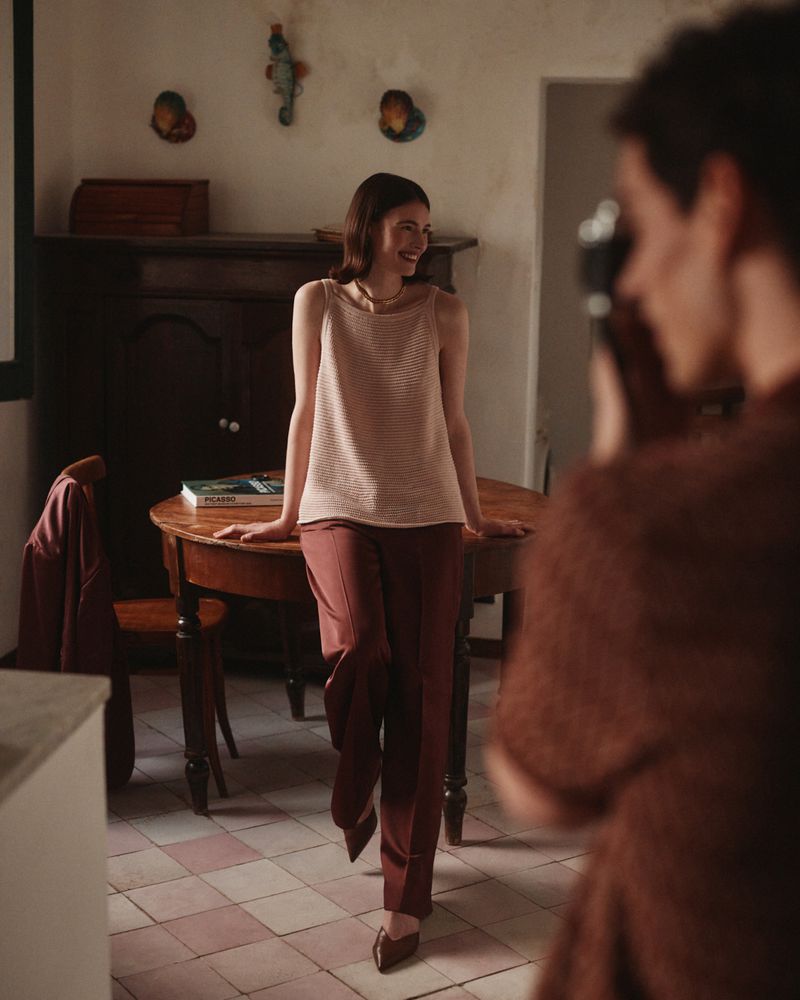 Smiling woman poses in casual top and pants indoors in a lifestyle shot as a photographer partially visible frames the picture.