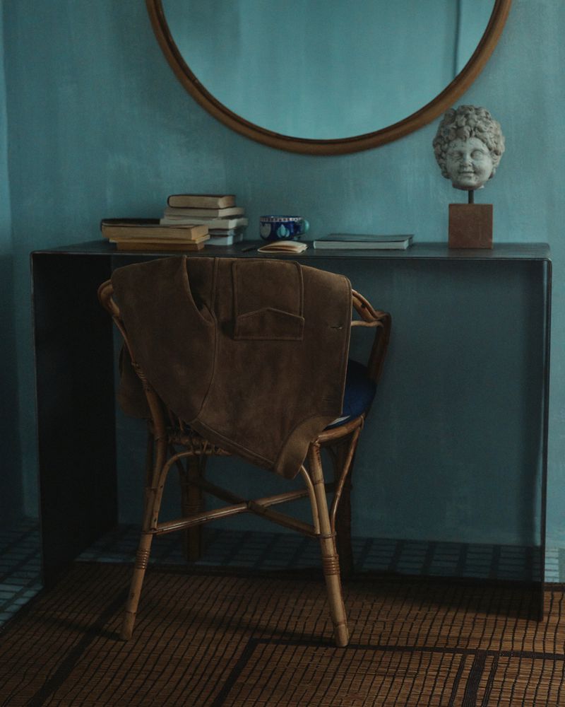 Stylish interior with a rattan chair, brown jacket, and a black console table with books and a bust, set against a teal wall.