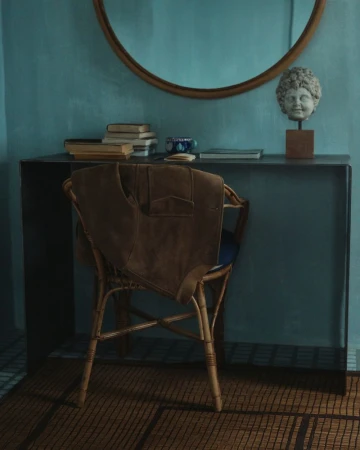 Stylish interior with a rattan chair, brown jacket, and a black console table with books and a bust, set against a teal wall.