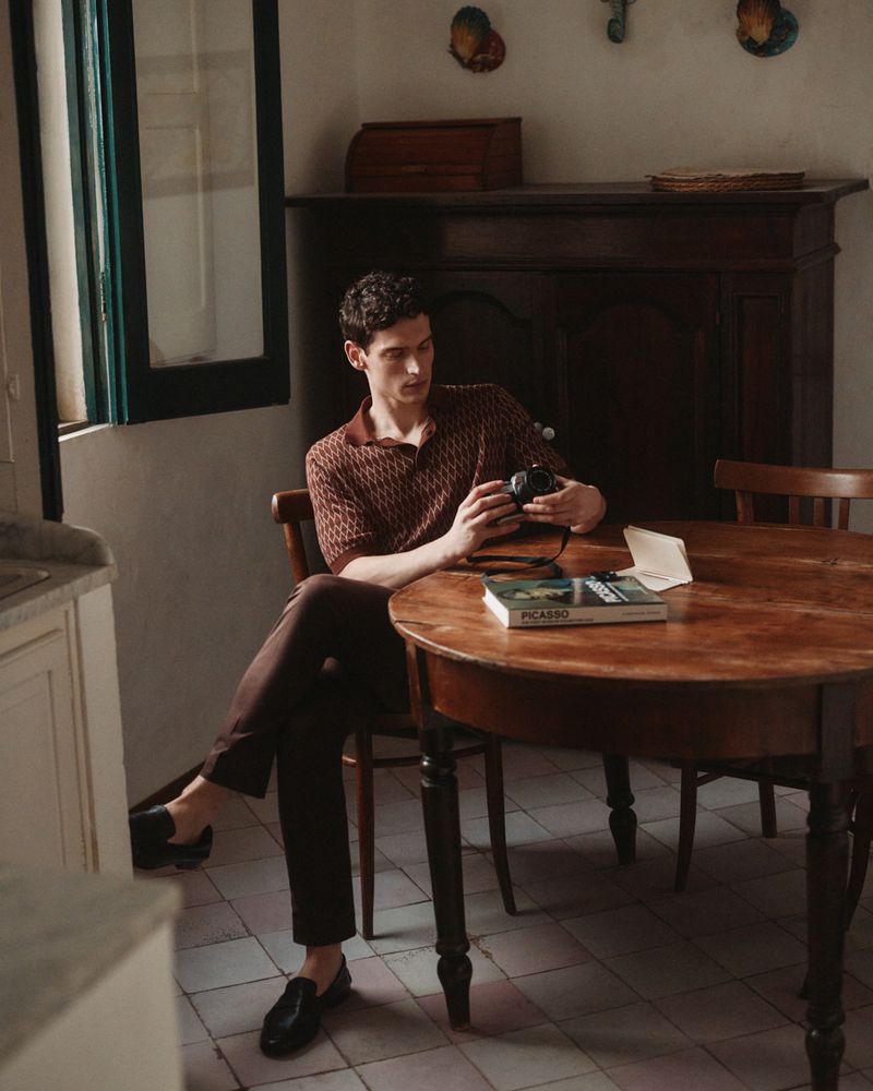 A man sits at a wooden table indoors, examining a camera. Sunlight streams in through a window. He's wearing a patterned shirt and dark pants.