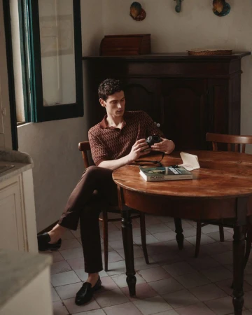 A man sits at a wooden table indoors, examining a camera. Sunlight streams in through a window. He's wearing a patterned shirt and dark pants.