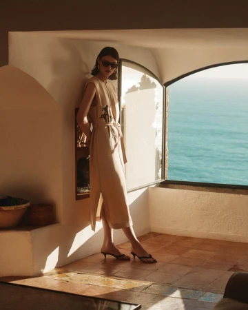 A slender woman in a cream-colored dress and sunglasses leans against a wall by a window with an ocean view in an indoor setting.