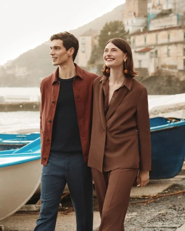 A stylish couple poses outdoors near boats and coastal buildings. The man wears an open rust shirt, while the woman sports a brown suit.