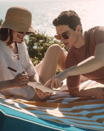 A man and woman relaxing on a striped beach towel outdoors in the bright sunlight, writing in a journal together.
