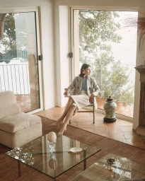Woman seated on a modern chair wearing a tweed blazer and cream skirt in a bright room with large windows