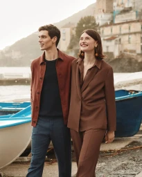 A man and woman standing outdoors near docked boats along a coast, wearing brown and navy apparel for Slowear SS26.