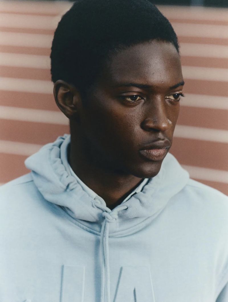 Portrait of a Black man wearing a light blue hoodie with the same color block-striped wall in the background in soft, diffused lighting.