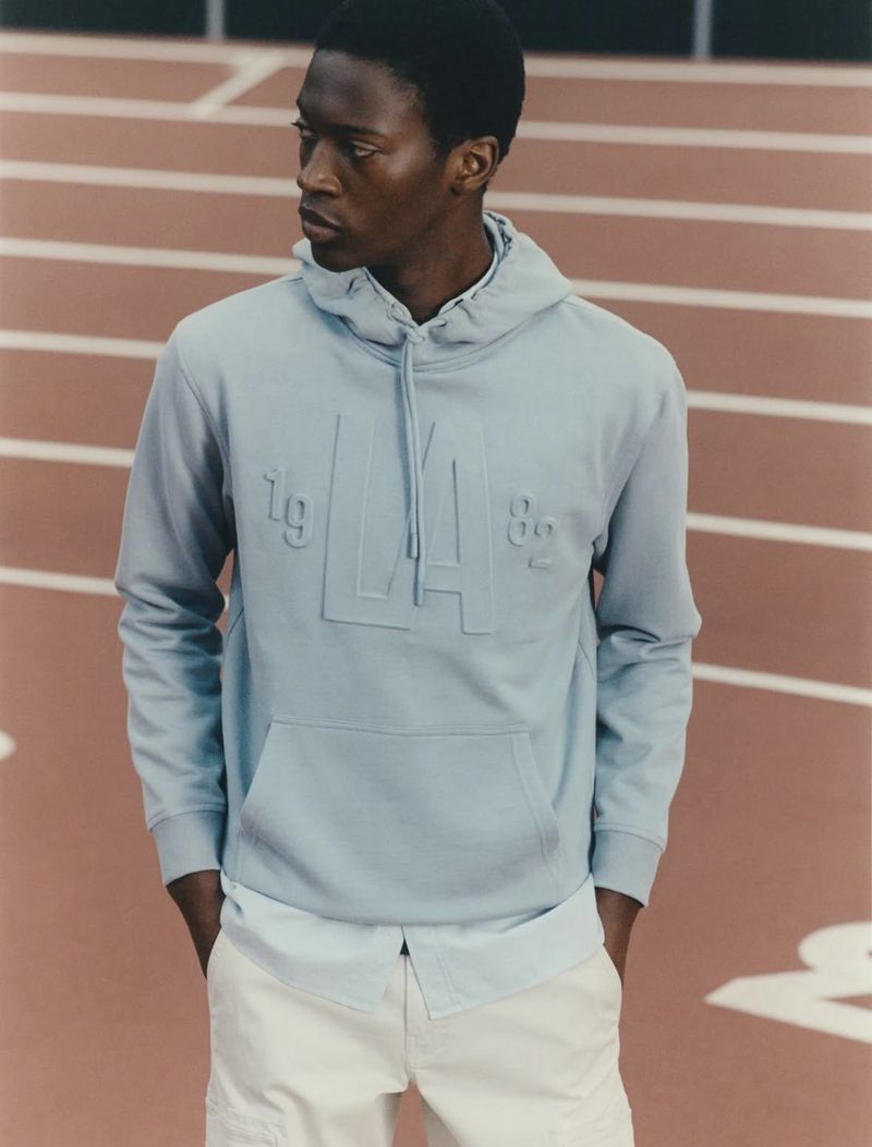 A Black man wearing a light blue hoodie with an 'LA 82' logo and light-colored pants poses against a blurred track background.