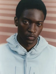 Close-up portrait of a Black man wearing a light blue hoodie against a slatted background for Sfera.