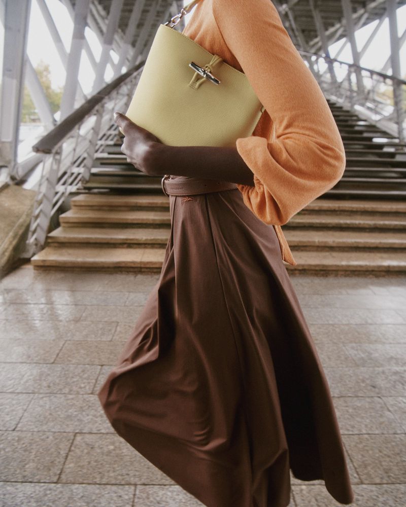 Woman in brown skirt walks beneath a gray metal structure holding a light green leather handbag with a chain strap.