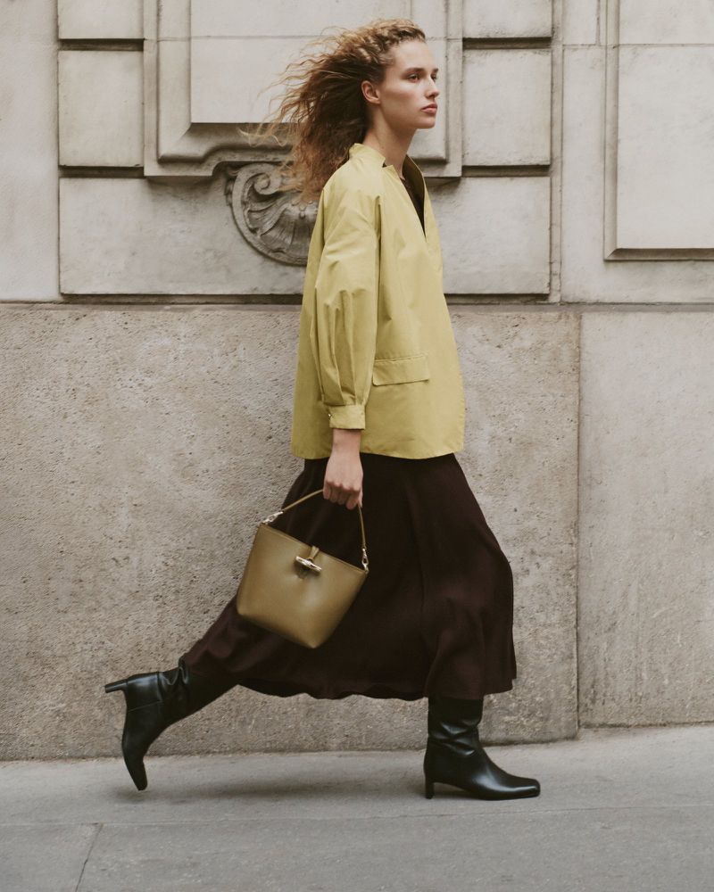 Woman in yellow jacket and brown skirt walks with handbag on city street. Fashion editorial photo with simple architectural background.