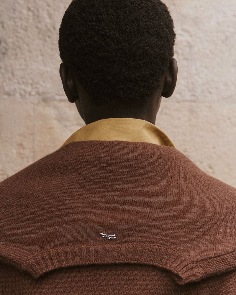 Back view of a person wearing a brown sweater with a mustard yellow collar, against a textured neutral colored wall.