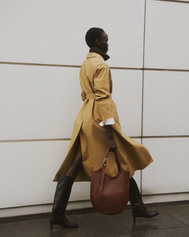 A stylish African American woman strides confidently in a tan trench coat, black tights, boots, carrying a brown leather hobo bag against a tiled wall.