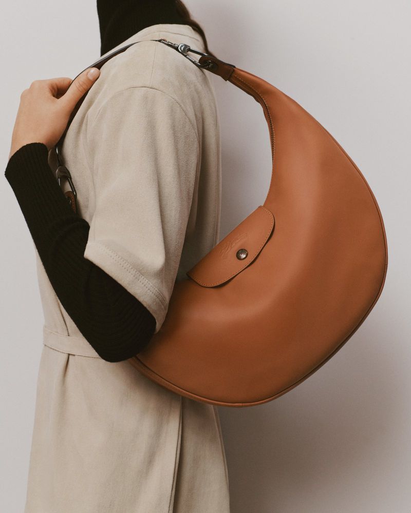 Person carrying a tan leather crescent handbag over their shoulder, wearing a black turtleneck and beige coat in a studio setting.