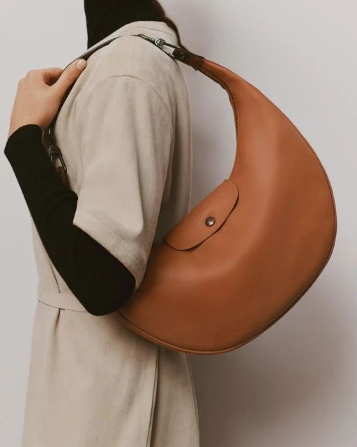Person carrying a tan leather crescent handbag over their shoulder, wearing a black turtleneck and beige coat in a studio setting.