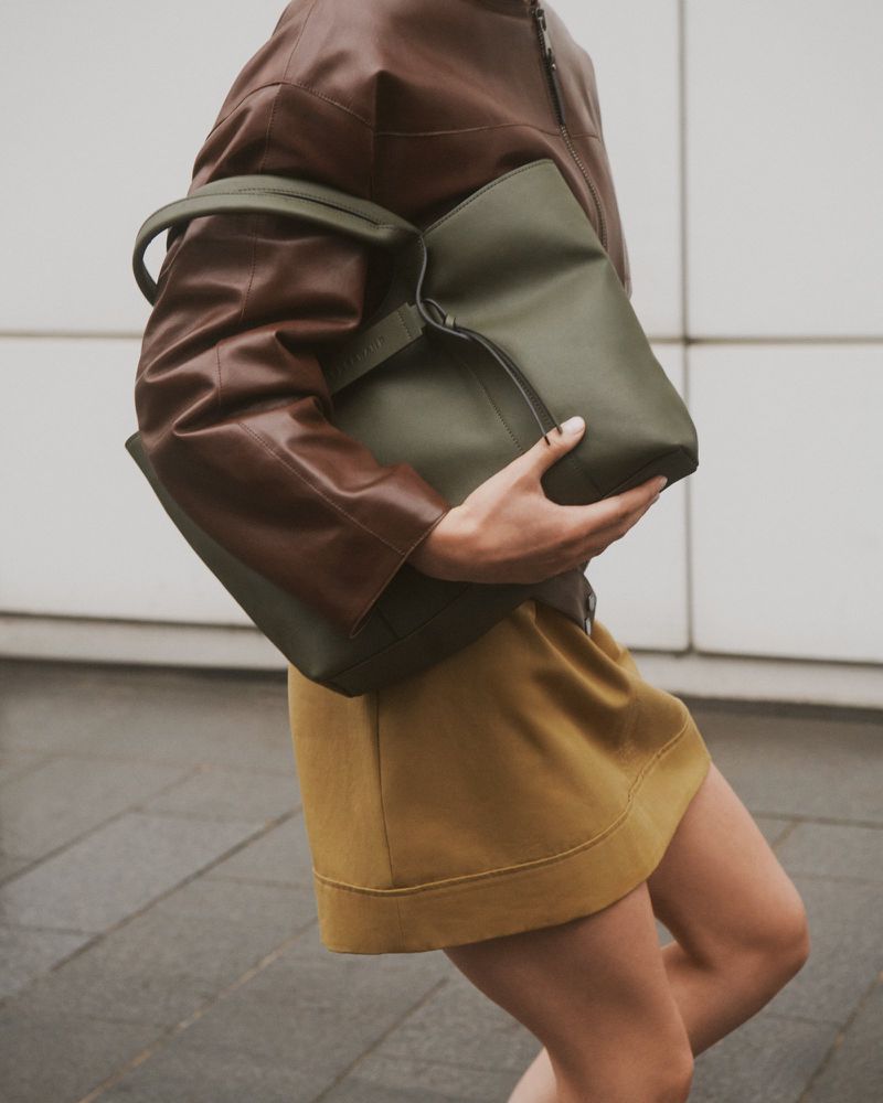 A woman carries a large green tote bag while walking outdoors, wearing a brown jacket and a mustard-colored skirt against a neutral wall.