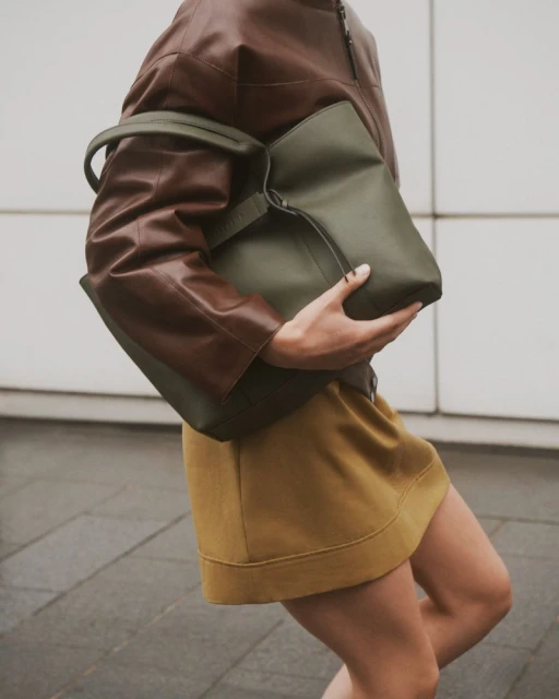 A woman carries a large green tote bag while walking outdoors, wearing a brown jacket and a mustard-colored skirt against a neutral wall.