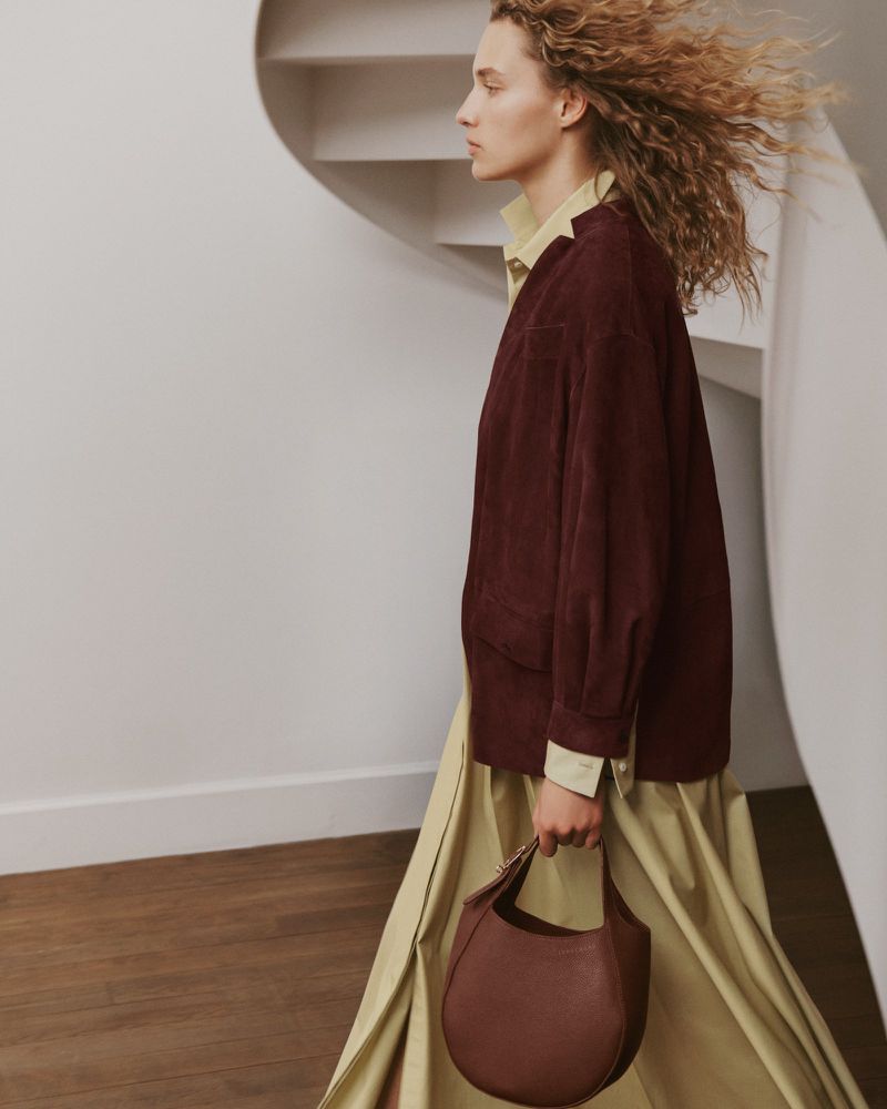 Fashionable woman with auburn handbag poses in front of a white stairway. She wears a chartreuse dress and a burgundy jacket.