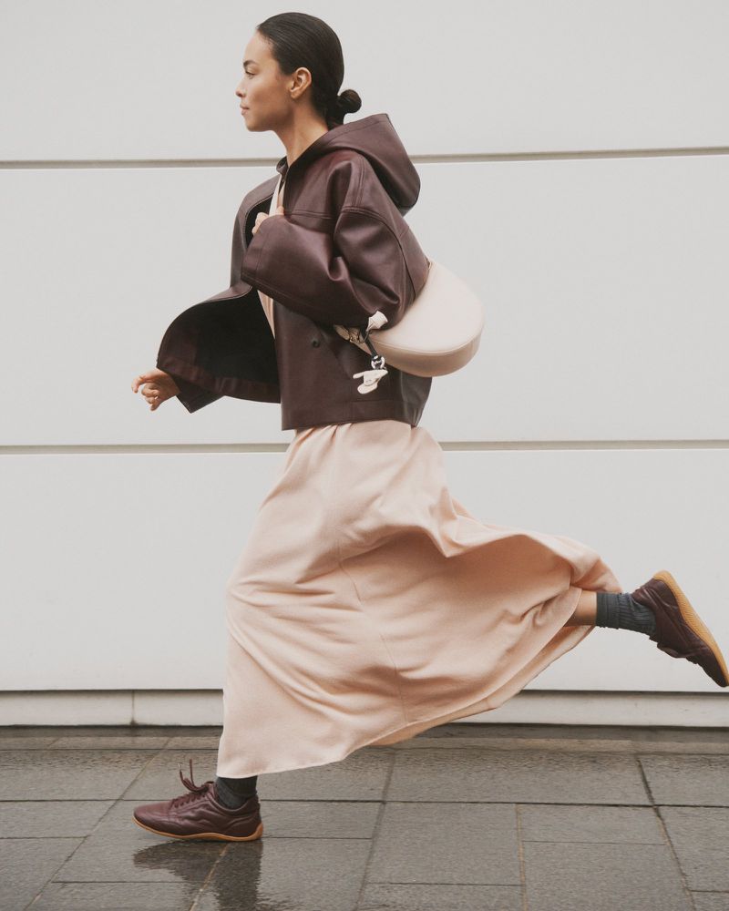 A woman is walking outdoors in a brown leather jacket, light pink skirt, and matching sneakers, carrying a light-colored purse over her shoulder.