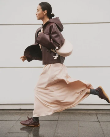A woman is walking outdoors in a brown leather jacket, light pink skirt, and matching sneakers, carrying a light-colored purse over her shoulder.
