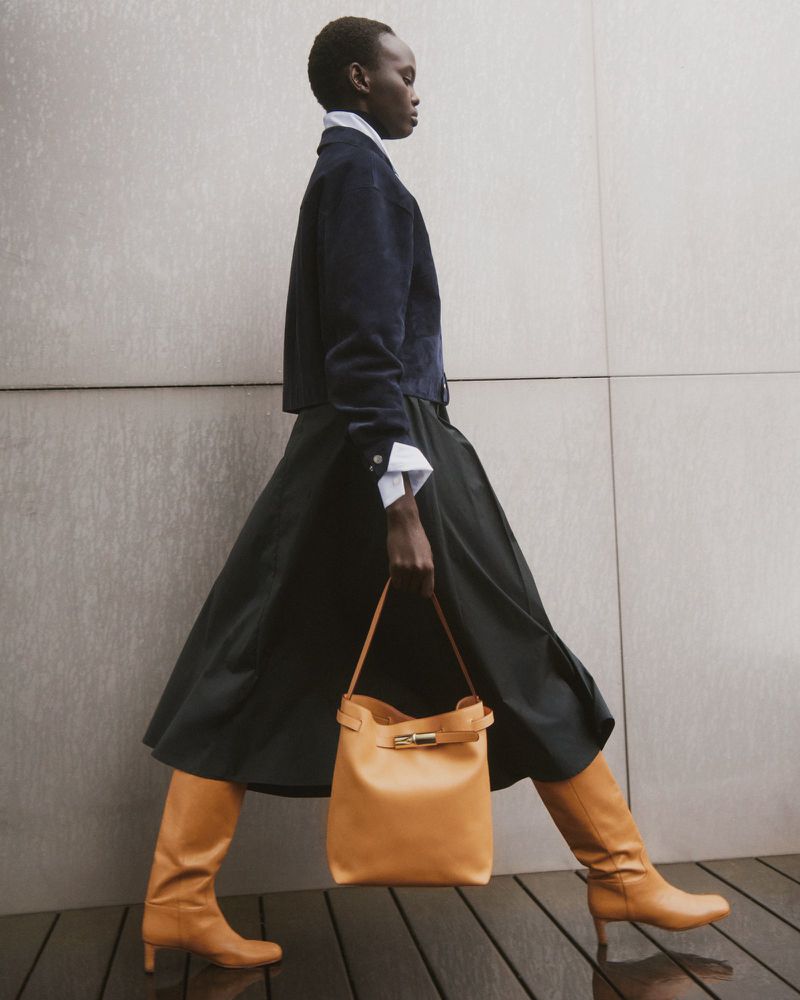 Full-length shot of an African female model in a navy jacket, black skirt, tan boots, and tan leather handbag on a plain background.