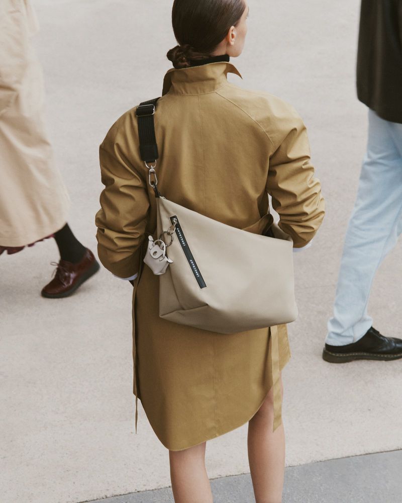 Back view of an adult female in a tan trench coat carrying a beige crossbody bag with people walking in the blurred background.