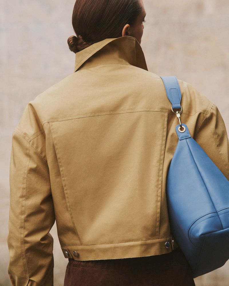 Back view of a woman wearing a tan jacket and carrying a blue leather handbag over her shoulder against a neutral wall.