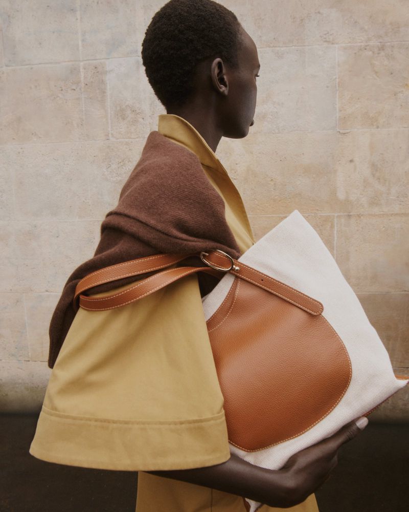 A woman with dark skin and short hair poses in a tan trench coat and brown shawl, holding a white tote bag with brown leather accents.