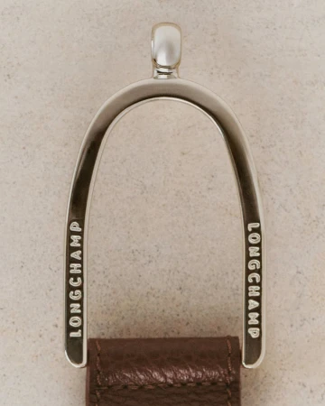 Close-up of a polished metal loop attached to a brown leather strap with the brand name Longchamp engraved on either side of the metal.