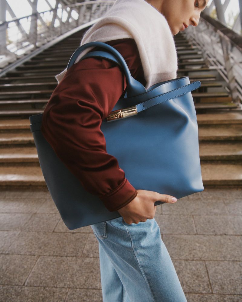 Woman wearing a scarf carries a blue leather tote bag and walks on an outdoor staircase with metal railings. The bag is the focus.