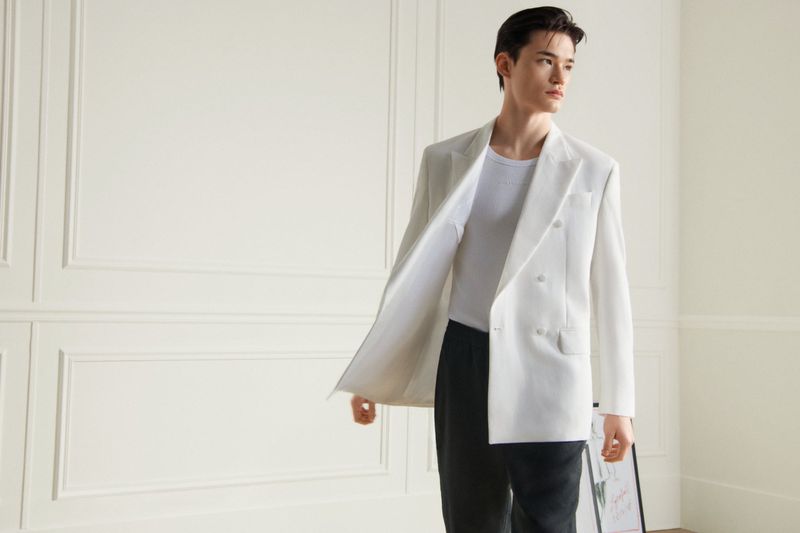 Elegant man in a white double-breasted blazer poses in a bright room with a paneled wall. Fashion editorial shot.
