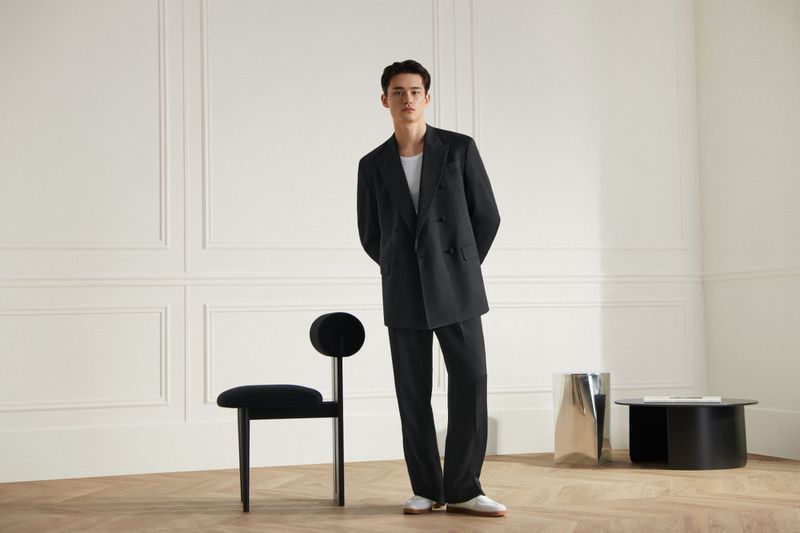 A young adult man posing in a black double-breasted suit in a minimalist interior setting, standing near a black chair and modern table.