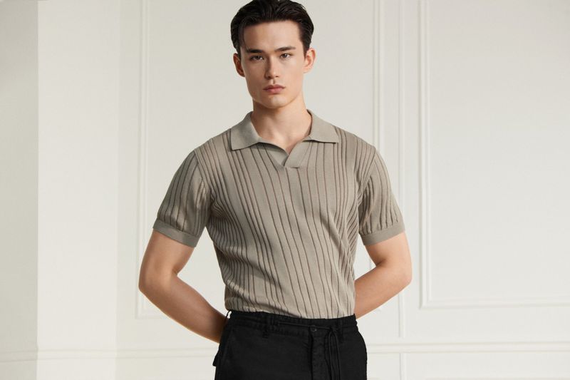 A stylish adult male in a ribbed knit polo shirt and black trousers, posing indoors with a relaxed expression. Fashion and portrait photography.